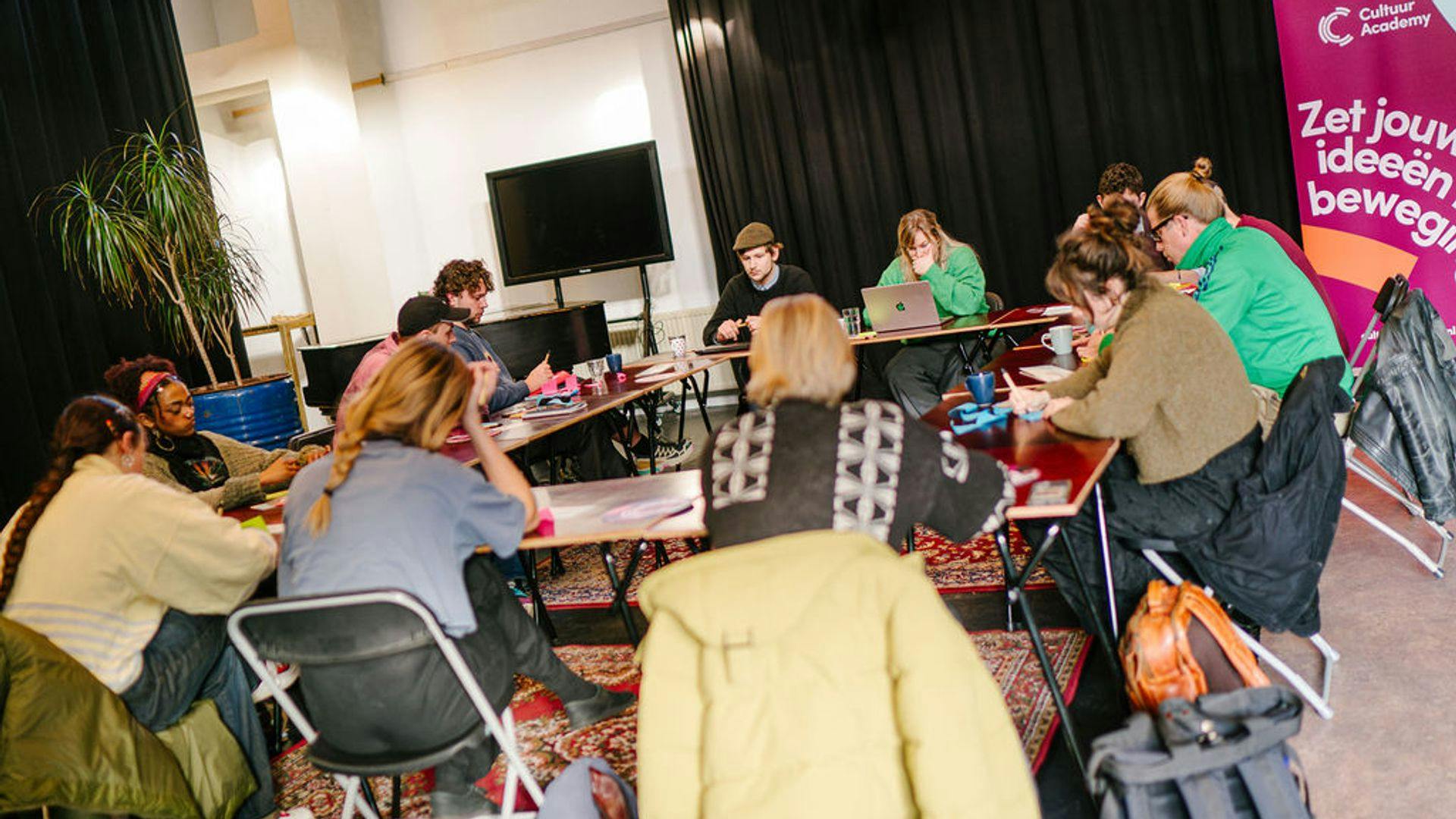 Foto van een grote groep jonge mensen die aan tafels werken bij een programma van Cultuur Academy.